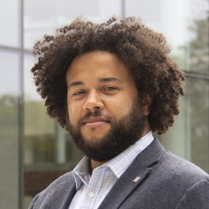 The image shows a man with curly brown hair and a beard. He is wearing a gray blazer over a checkered shirt. The background appears to be a modern building with glass windows. The man is looking directly at the camera with a slight smile.